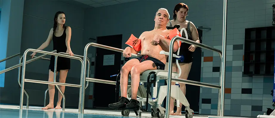 En rullstolsburen man är på väg ner i rehabbassängen med hjälp av två kvinnor. 